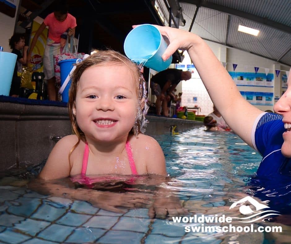 toddler smiling in water with swim teaching next to her holding cup of water and demonstrating water pouring over head