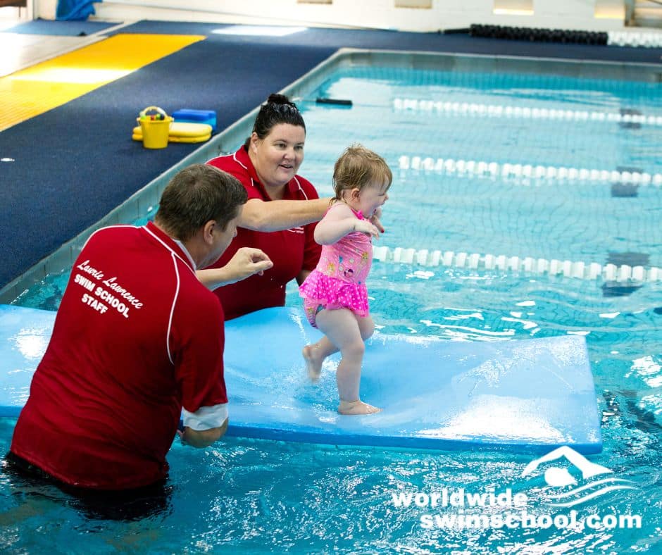 toddler walking on mat in the water with two teachers holding child's hands