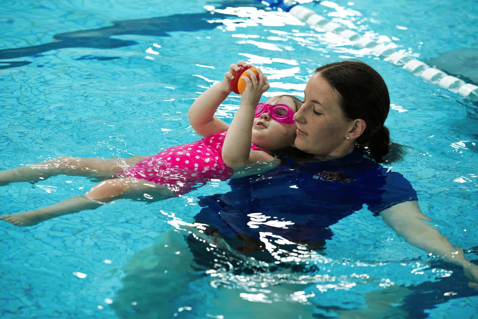 Swim teacher with infant floating on back with ball in hands looking up 